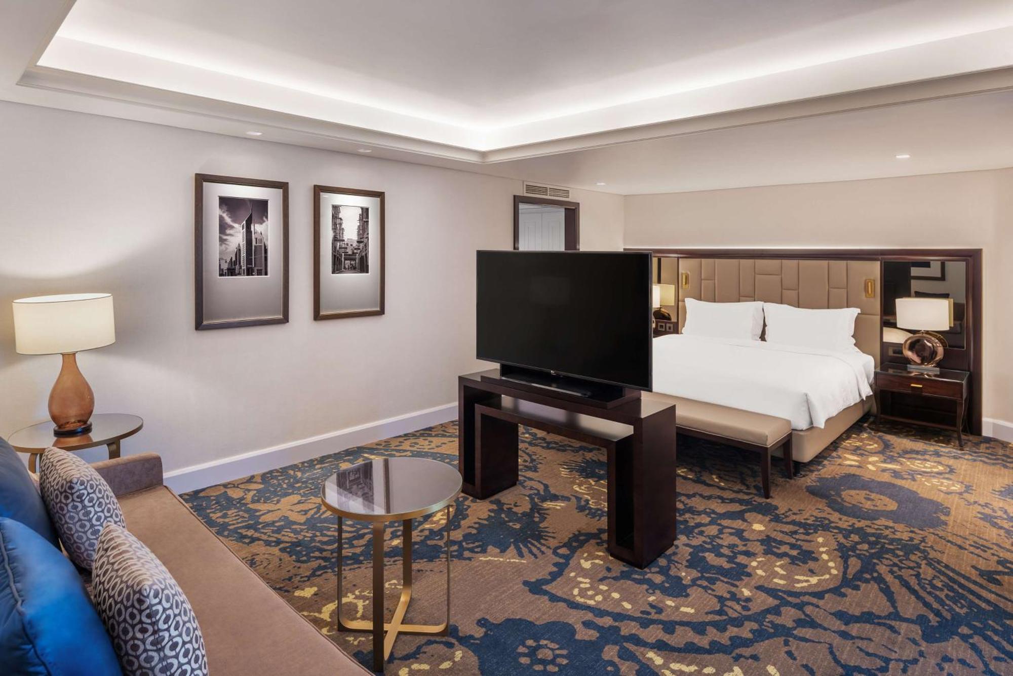 Radisson Blu Hotel, Dubai Deira Creek Esterno foto The image shows an elegant hotel room. It features a neutral color palette with modern furnishings. On one side, there is a comfortable sofa and a small round table. A large flat-screen TV is positioned on a dark wooden stand in the center. The room 