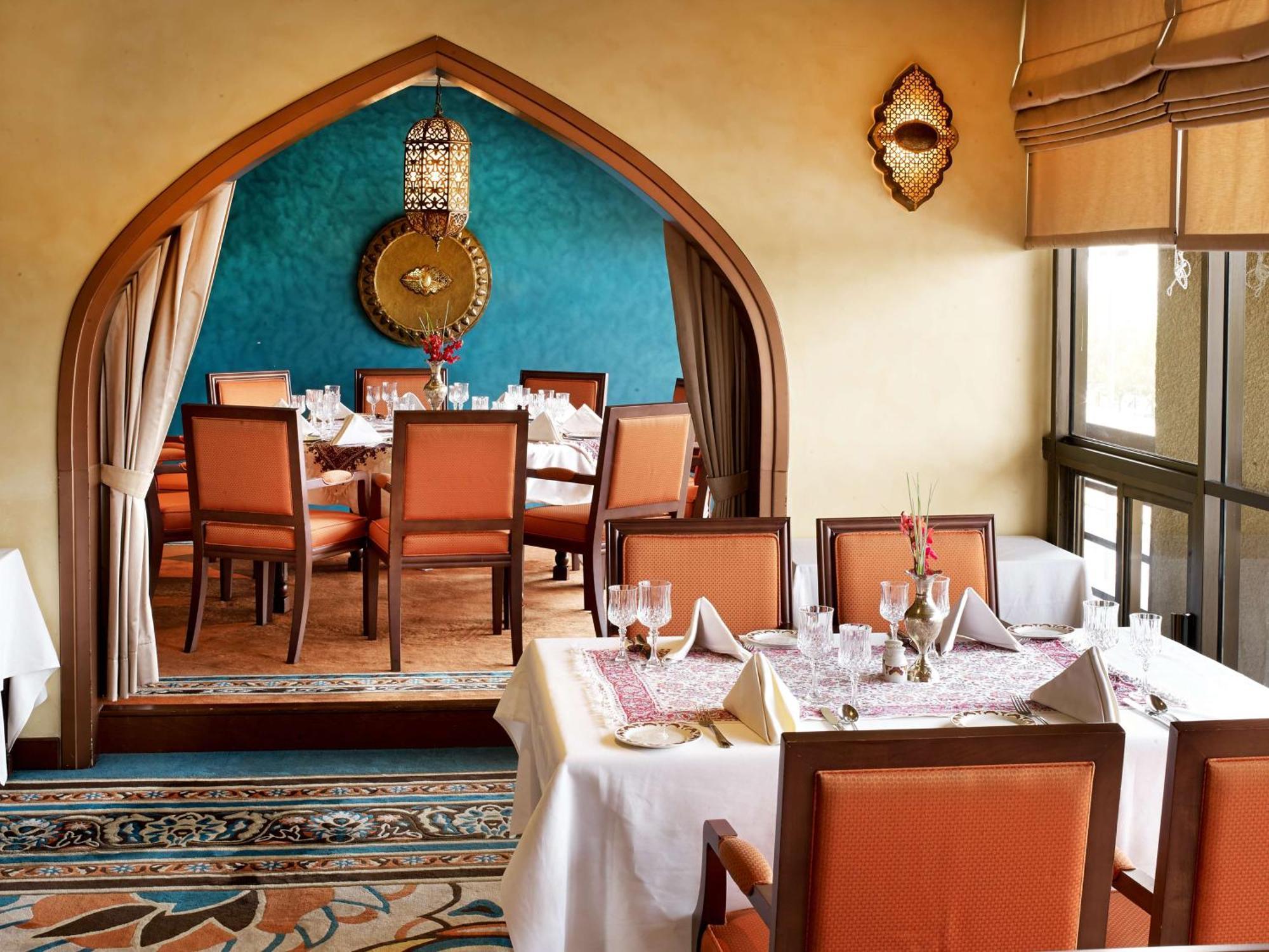 Radisson Blu Hotel, Dubai Deira Creek Esterno foto The photo shows a restaurant interior with a warm and inviting ambiance. In the foreground, there are elegantly set tables covered with white tablecloths and neat place settings, including folded napkins. The background features an archway leading in