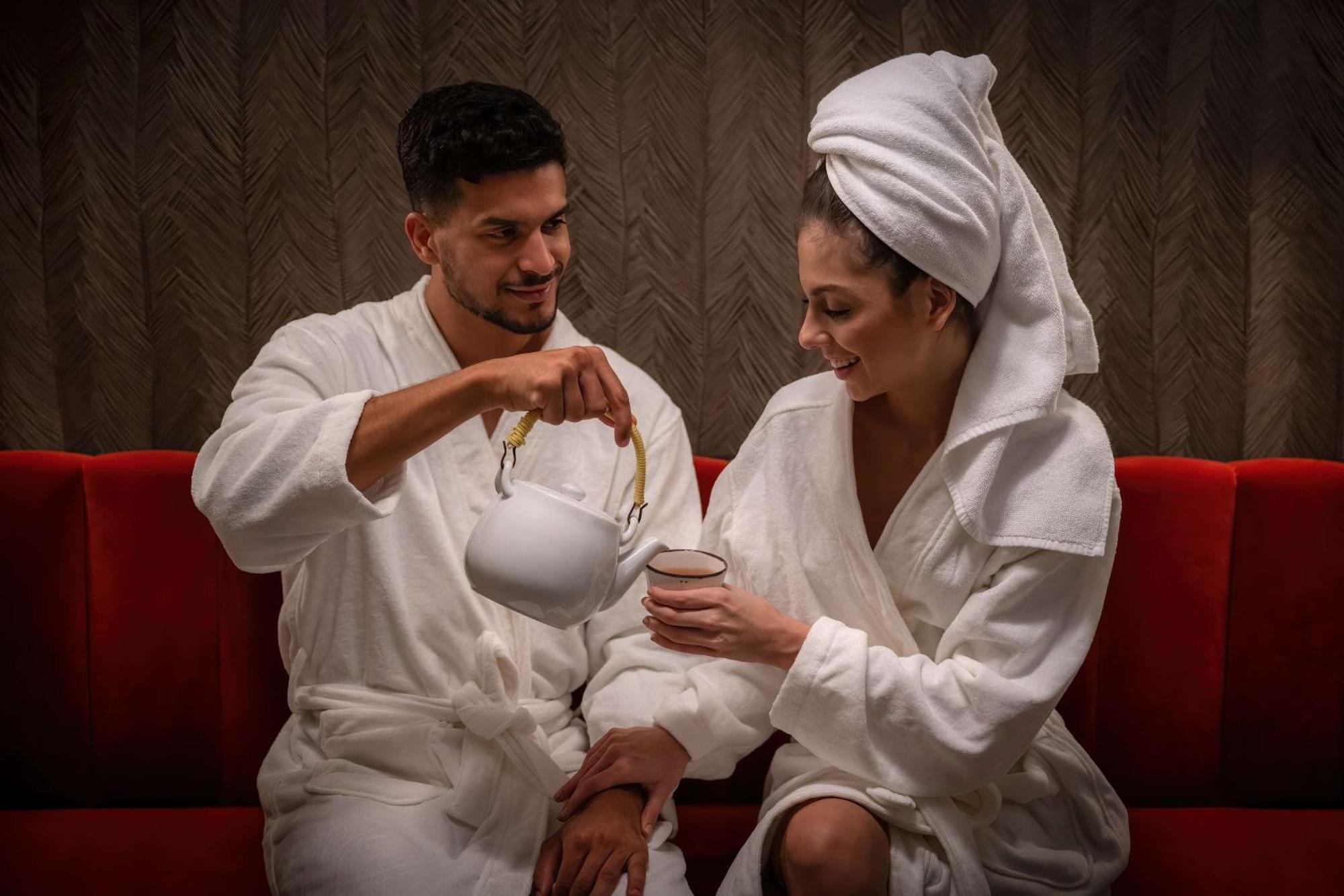 Radisson Blu Hotel, Dubai Deira Creek Esterno foto The photo depicts a man and a woman sitting together on a red couch, both dressed in white bathrobes. The man is pouring tea from a white teapot into a cup held by the woman, who is smiling. She has a towel wrapped around her head, suggesting a relax