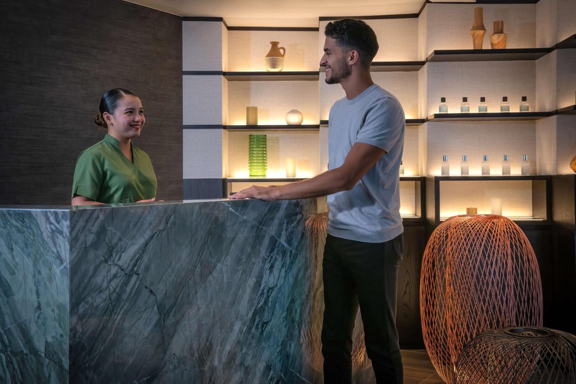 Radisson Blu Hotel, Dubai Deira Creek Esterno foto The image shows a reception area, likely in a spa or wellness center. On one side, there is a woman wearing a green uniform, smiling at a man who is standing at a marble reception desk. The man is dressed casually in a gray t-shirt and dark pants. Be